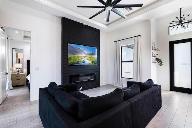 living room with light hardwood / wood-style floors, ceiling fan with notable chandelier, a fireplace, a raised ceiling, and ornamental molding