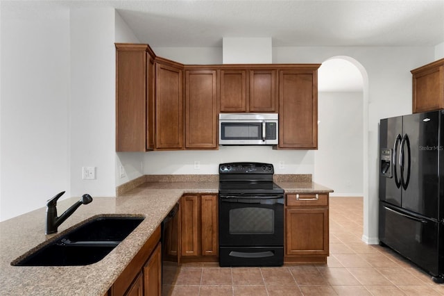 kitchen featuring arched walkways, brown cabinetry, a sink, light stone countertops, and black appliances