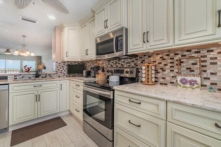 kitchen with cream cabinets, hanging light fixtures, backsplash, appliances with stainless steel finishes, and light stone counters