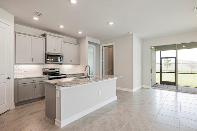 kitchen with gray cabinetry, a center island with sink, sink, appliances with stainless steel finishes, and light stone counters