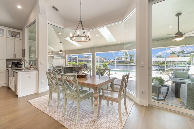 dining space with lofted ceiling with skylight, ceiling fan with notable chandelier, a water view, and light wood-type flooring