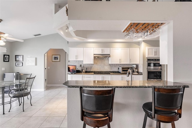 kitchen with dark stone countertops, double oven, a kitchen breakfast bar, and white cabinets