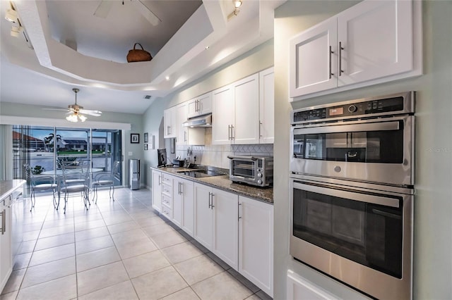 kitchen featuring light tile patterned flooring, dark stone countertops, a tray ceiling, stainless steel double oven, and white cabinets