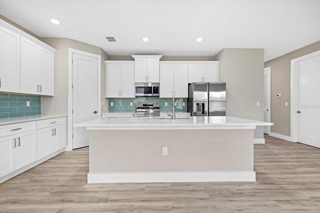 kitchen with appliances with stainless steel finishes, backsplash, a center island with sink, light hardwood / wood-style flooring, and white cabinets