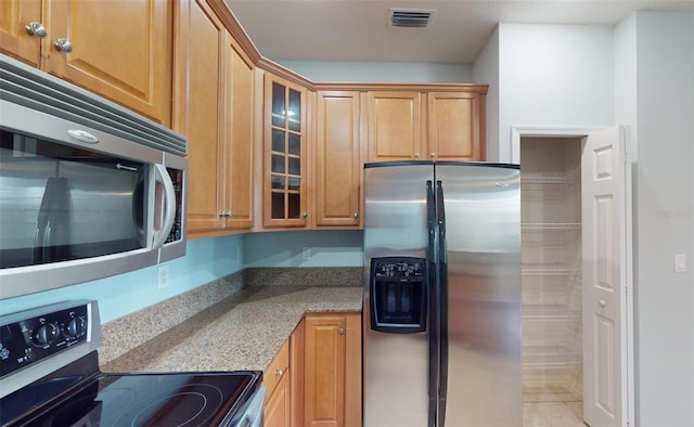 kitchen featuring light stone counters, brown cabinets, glass insert cabinets, appliances with stainless steel finishes, and visible vents