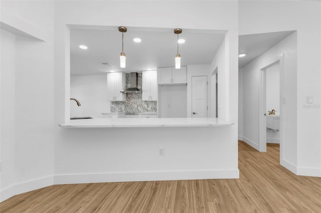 kitchen with white cabinetry, wall chimney range hood, hanging light fixtures, kitchen peninsula, and light hardwood / wood-style flooring