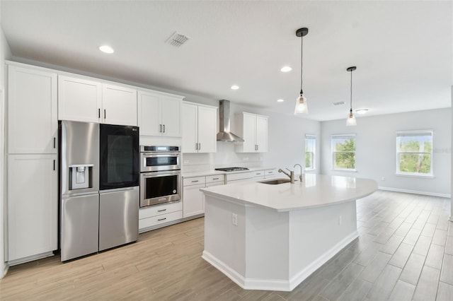 kitchen with stainless steel appliances, white cabinetry, wall chimney exhaust hood, an island with sink, and pendant lighting