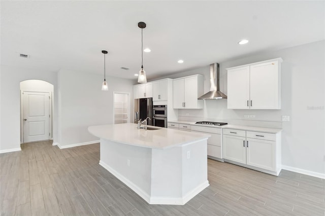 kitchen with stainless steel appliances, a kitchen island with sink, wall chimney range hood, white cabinetry, and decorative light fixtures