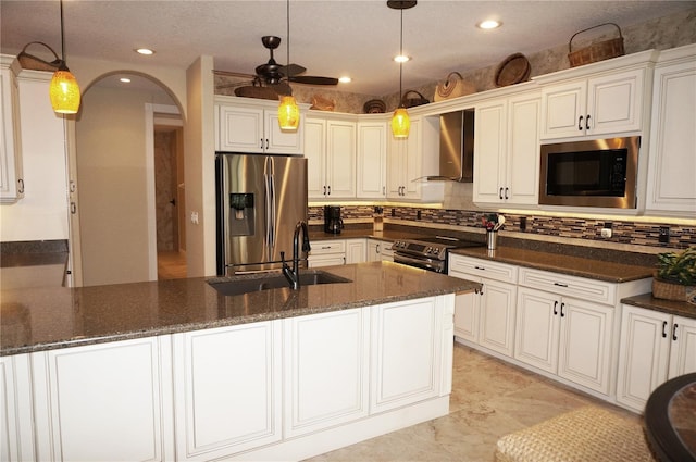 kitchen featuring stainless steel appliances, wall chimney exhaust hood, a sink, dark stone counters, and tasteful backsplash