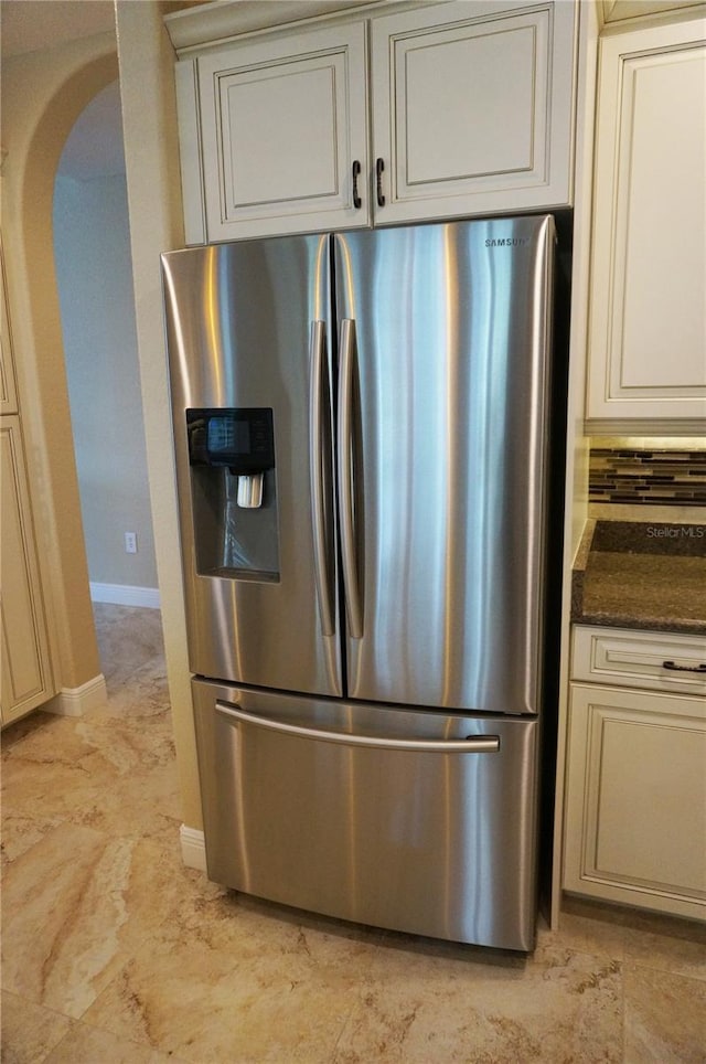 kitchen with stainless steel refrigerator with ice dispenser, arched walkways, baseboards, cream cabinets, and dark stone counters