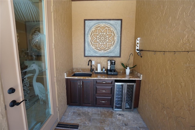 bar with light stone counters, sink, beverage cooler, and dark brown cabinets