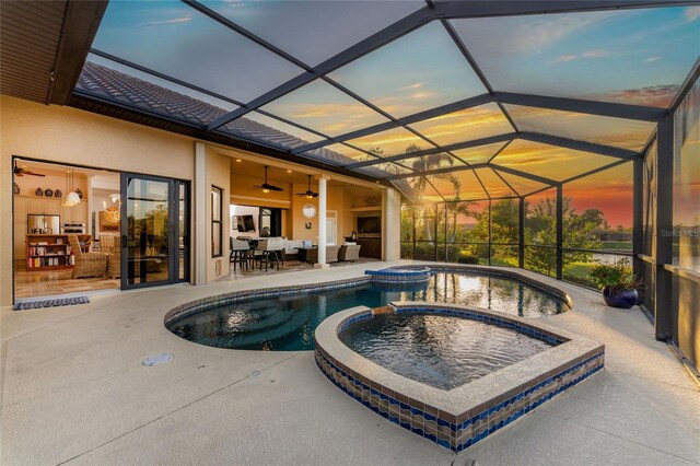 pool at dusk featuring an in ground hot tub, glass enclosure, ceiling fan, and a patio area