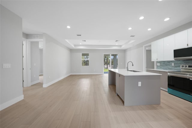 kitchen featuring sink, appliances with stainless steel finishes, a tray ceiling, an island with sink, and light wood-type flooring