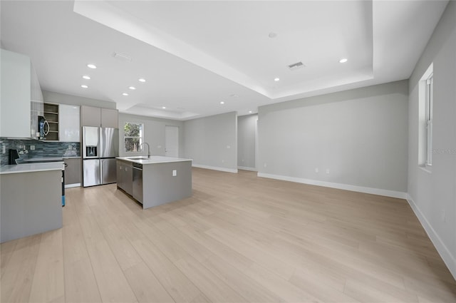 kitchen featuring stainless steel appliances, tasteful backsplash, an island with sink, a tray ceiling, and light hardwood / wood-style flooring