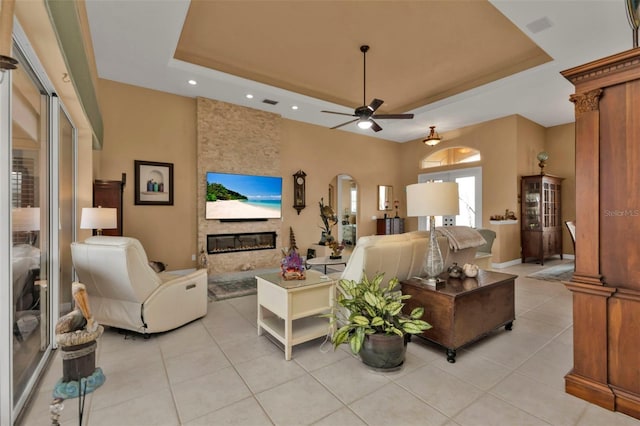 tiled living room featuring a stone fireplace, ceiling fan, and a tray ceiling