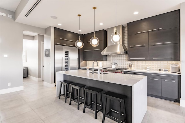 kitchen featuring sink, wall chimney exhaust hood, backsplash, pendant lighting, and a center island with sink