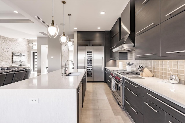 kitchen featuring high end appliances, wall chimney range hood, sink, an island with sink, and decorative light fixtures