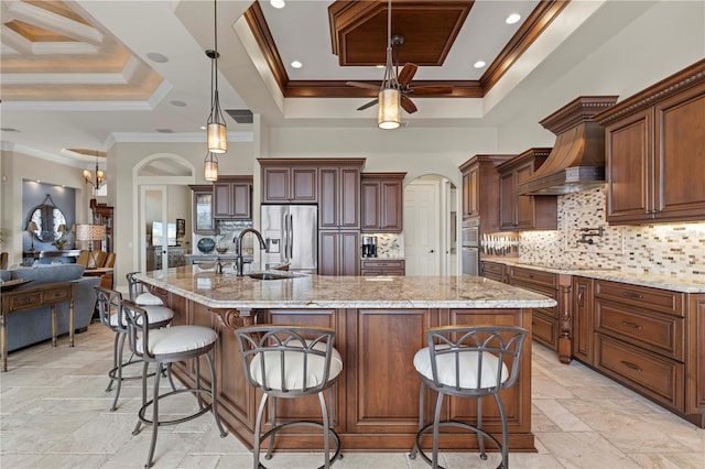 kitchen with premium range hood, a large island with sink, stainless steel fridge, a tray ceiling, and ornamental molding