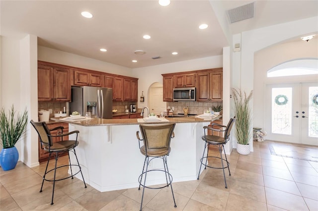 kitchen with a kitchen island with sink, french doors, light stone countertops, appliances with stainless steel finishes, and a breakfast bar area