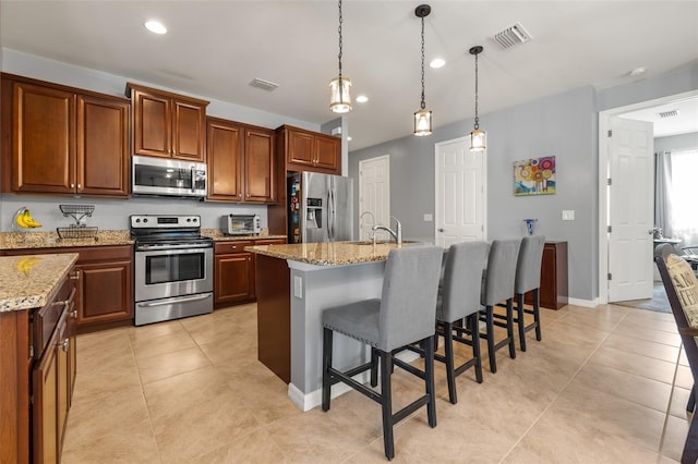 kitchen featuring a breakfast bar, appliances with stainless steel finishes, light stone counters, an island with sink, and decorative light fixtures