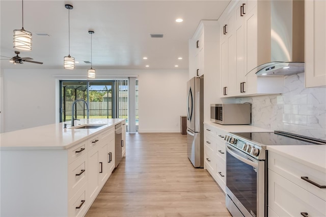 kitchen with appliances with stainless steel finishes, a kitchen island with sink, wall chimney range hood, decorative light fixtures, and white cabinetry