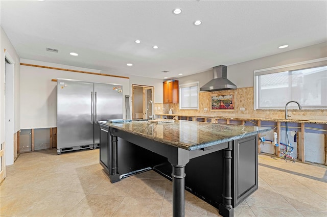 kitchen featuring a large island with sink, high end fridge, dark stone countertops, and wall chimney range hood