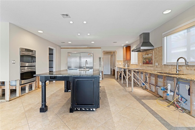 kitchen featuring a center island with sink, wall chimney exhaust hood, stone countertops, a kitchen bar, and stainless steel appliances