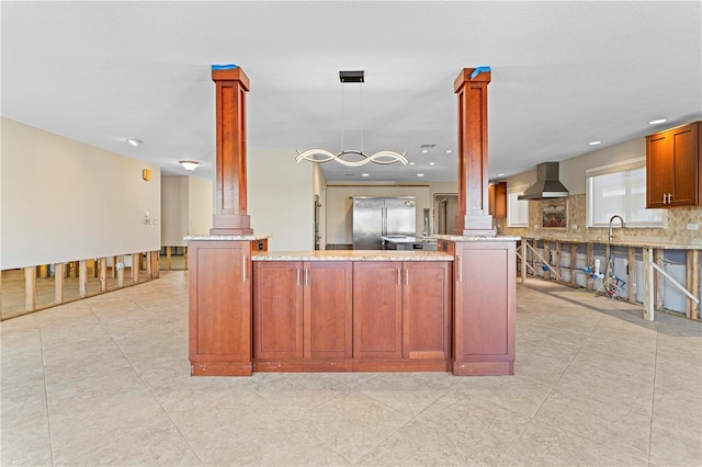 kitchen featuring kitchen peninsula, decorative backsplash, decorative columns, wall chimney exhaust hood, and stainless steel refrigerator