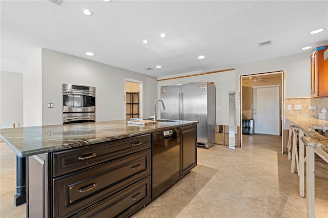 kitchen with dark stone counters, dark brown cabinetry, stainless steel appliances, sink, and an island with sink