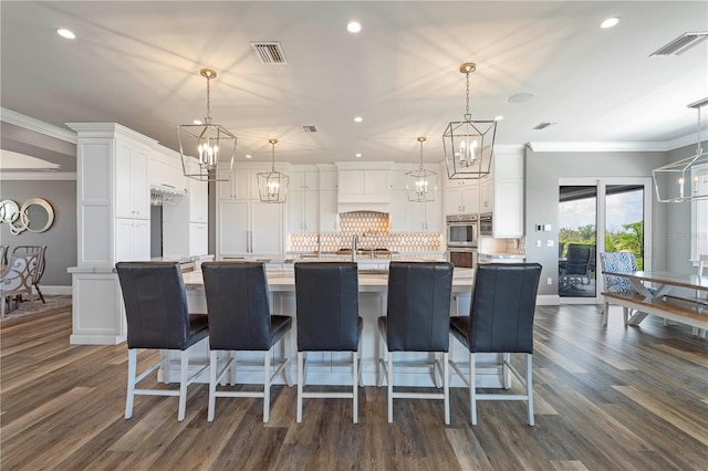 kitchen with ornamental molding, dark hardwood / wood-style floors, white cabinetry, hanging light fixtures, and an island with sink