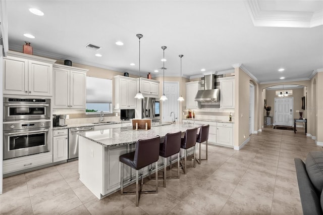 kitchen featuring ornamental molding, wall chimney exhaust hood, stainless steel appliances, sink, and an island with sink