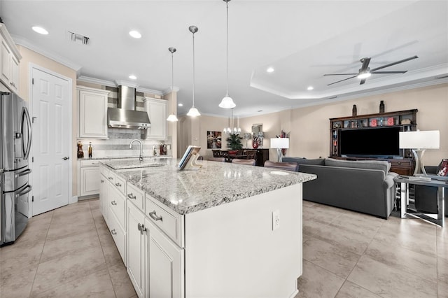 kitchen with pendant lighting, a kitchen island with sink, stainless steel refrigerator with ice dispenser, wall chimney exhaust hood, and white cabinetry