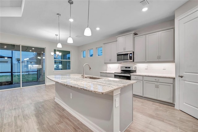 kitchen featuring a kitchen island with sink, sink, appliances with stainless steel finishes, decorative light fixtures, and light hardwood / wood-style floors