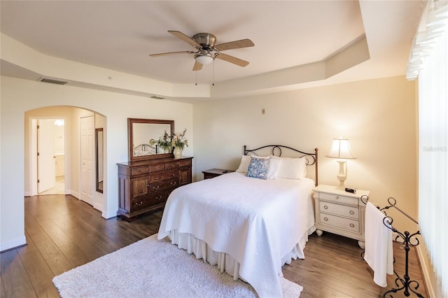 bedroom featuring a tray ceiling, ceiling fan, ensuite bathroom, and dark wood-type flooring