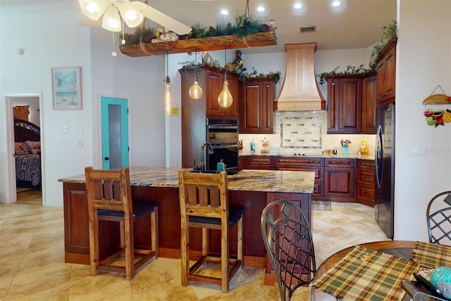 kitchen featuring a kitchen island with sink, stainless steel appliances, light stone counters, and custom exhaust hood