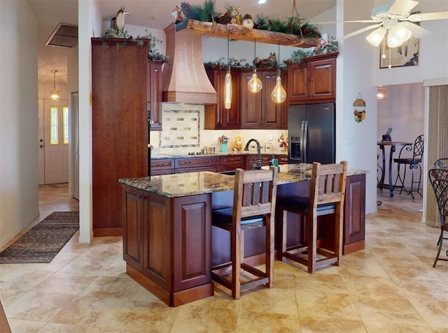 kitchen with light stone counters, high vaulted ceiling, stainless steel refrigerator with ice dispenser, a center island with sink, and custom exhaust hood