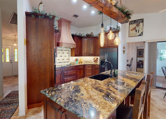 kitchen with custom exhaust hood, a kitchen island with sink, sink, dark stone countertops, and stainless steel fridge with ice dispenser