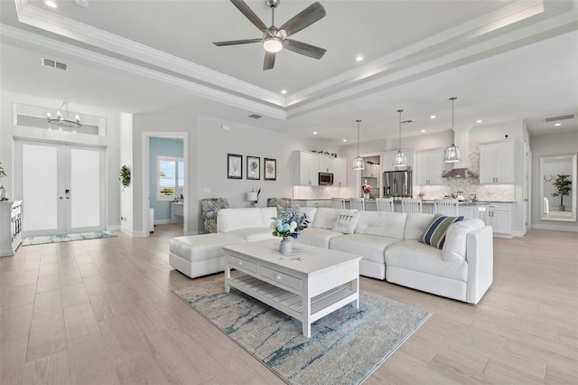 living room featuring french doors, ceiling fan with notable chandelier, a raised ceiling, crown molding, and light hardwood / wood-style floors