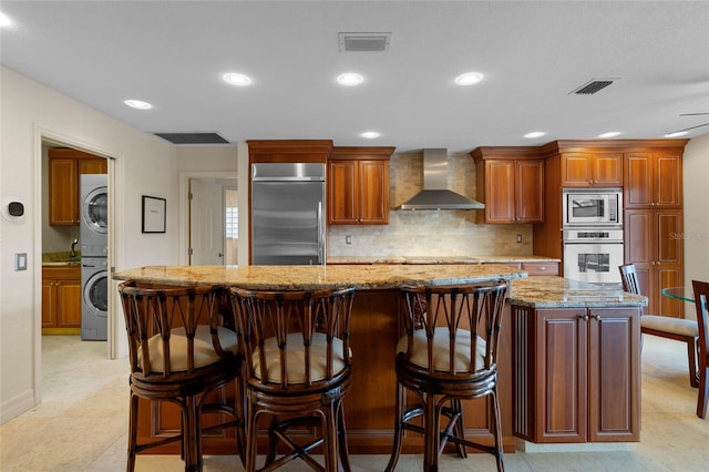 kitchen with wall chimney range hood, light stone counters, built in appliances, a kitchen island, and stacked washer and clothes dryer