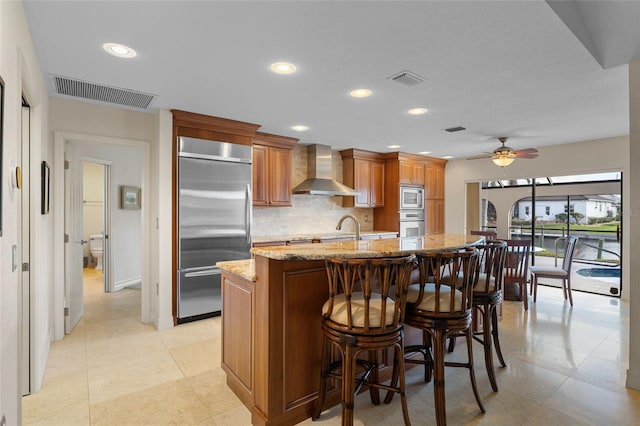 kitchen featuring light stone countertops, wall chimney exhaust hood, a breakfast bar, ceiling fan, and built in appliances