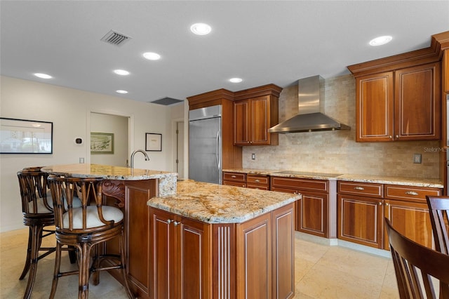 kitchen with light stone countertops, decorative backsplash, wall chimney range hood, built in fridge, and an island with sink