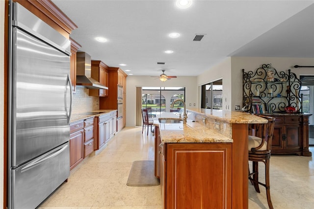 kitchen with sink, wall chimney range hood, a kitchen bar, stainless steel built in fridge, and a center island with sink