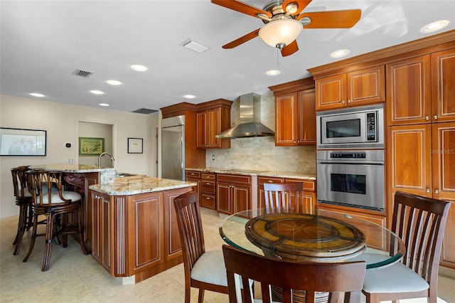 kitchen with light stone countertops, sink, wall chimney exhaust hood, built in appliances, and an island with sink