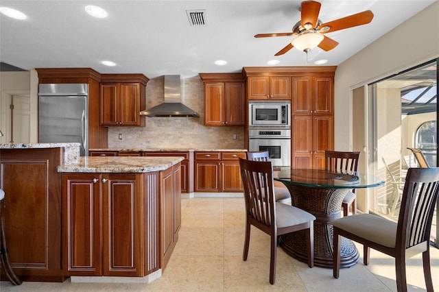 kitchen with wall chimney range hood, built in appliances, ceiling fan, light stone countertops, and tasteful backsplash