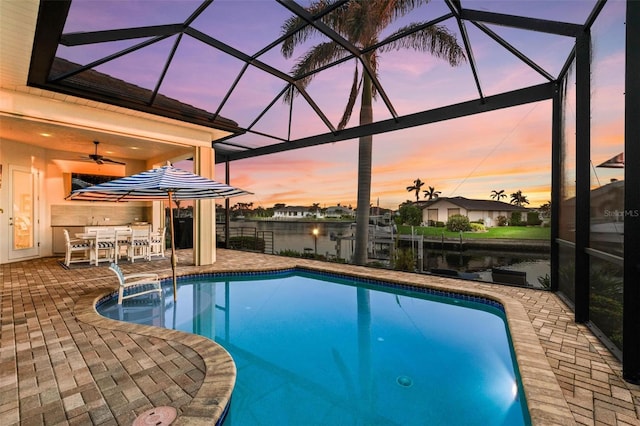 pool at dusk with a patio area, a water view, an outdoor bar, and glass enclosure