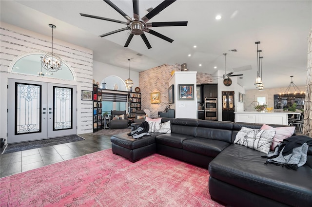 tiled living room featuring brick wall, ceiling fan with notable chandelier, french doors, and high vaulted ceiling