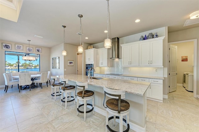 kitchen with white cabinets, wall chimney exhaust hood, an island with sink, and hanging light fixtures