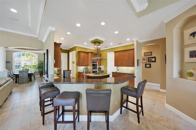 kitchen with an inviting chandelier, dark stone countertops, ornamental molding, island range hood, and a breakfast bar area