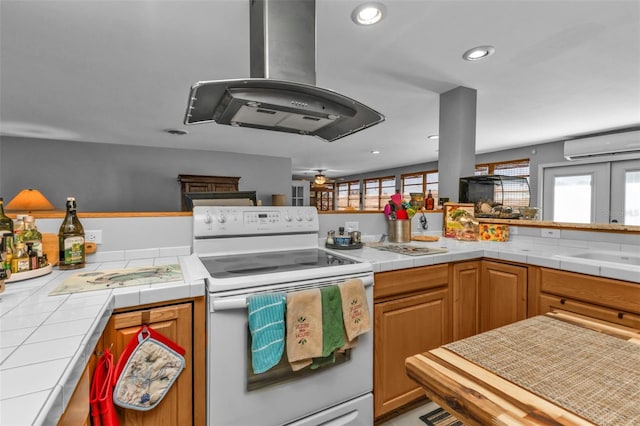 kitchen with tile counters, a wall mounted air conditioner, white range with electric stovetop, island exhaust hood, and kitchen peninsula