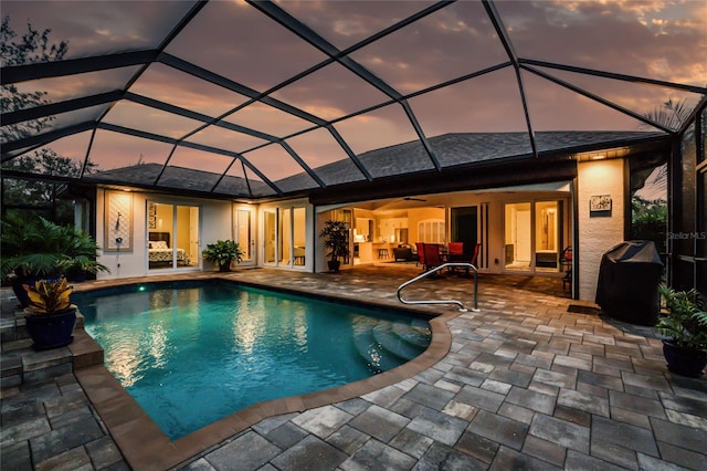 pool at dusk featuring area for grilling, glass enclosure, ceiling fan, and a patio area